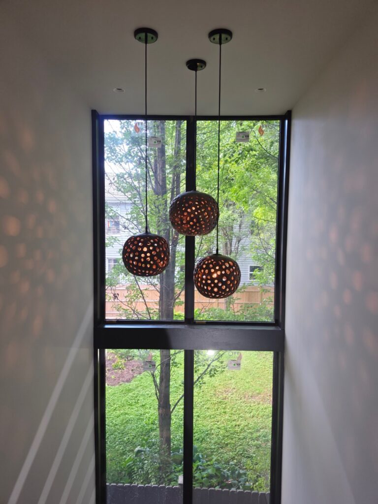 Three round, perforated metal pendant lights hanging in a stairwell in front of tall black-framed windows overlooking greenery