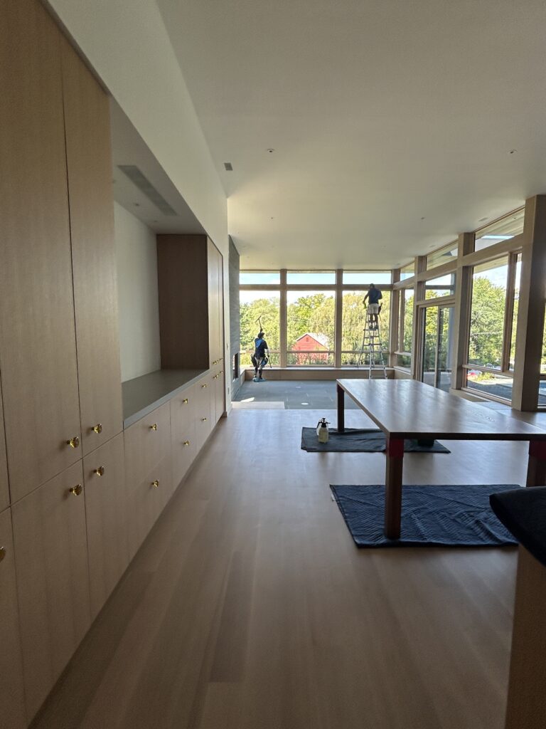 Bright open-concept living and dining room with light wood floors and floor-to-ceiling windows; workers cleaning tall corner windows overlooking trees and a red barn