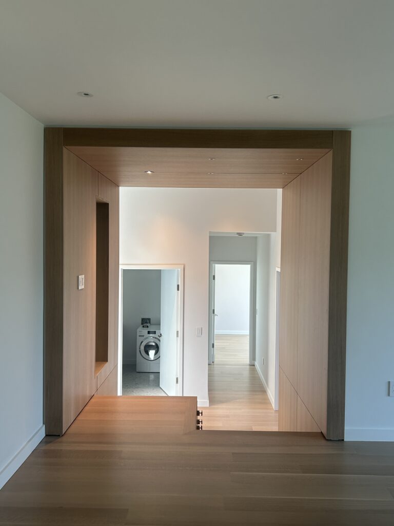Interior view through a light-wood-clad archway framing a hallway, with an open laundry room and washing machine visible ahead