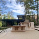 Small wood architectural model of a modern two-story home on a cardboard site base, held in front of the finished dark-clad house among trees.