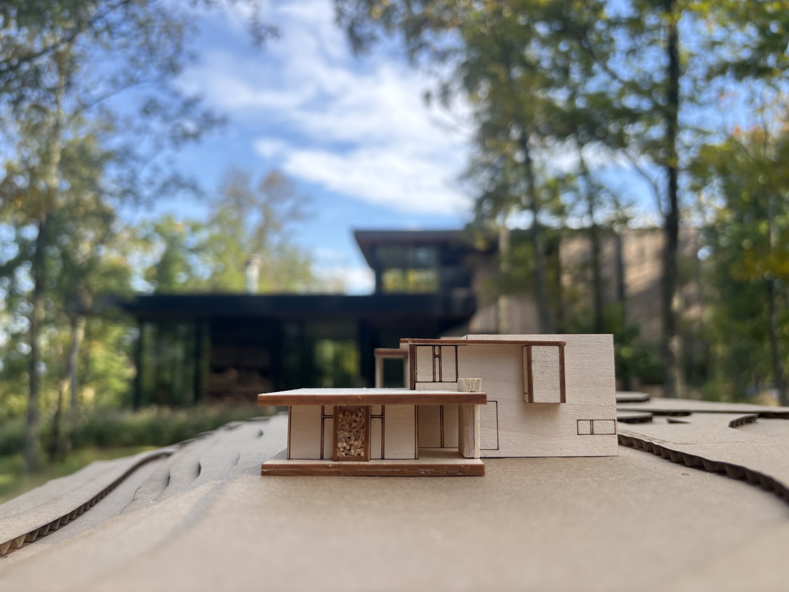 Small wood architectural model of a modern two-story home on a cardboard site base, held in front of the finished dark-clad house among trees.