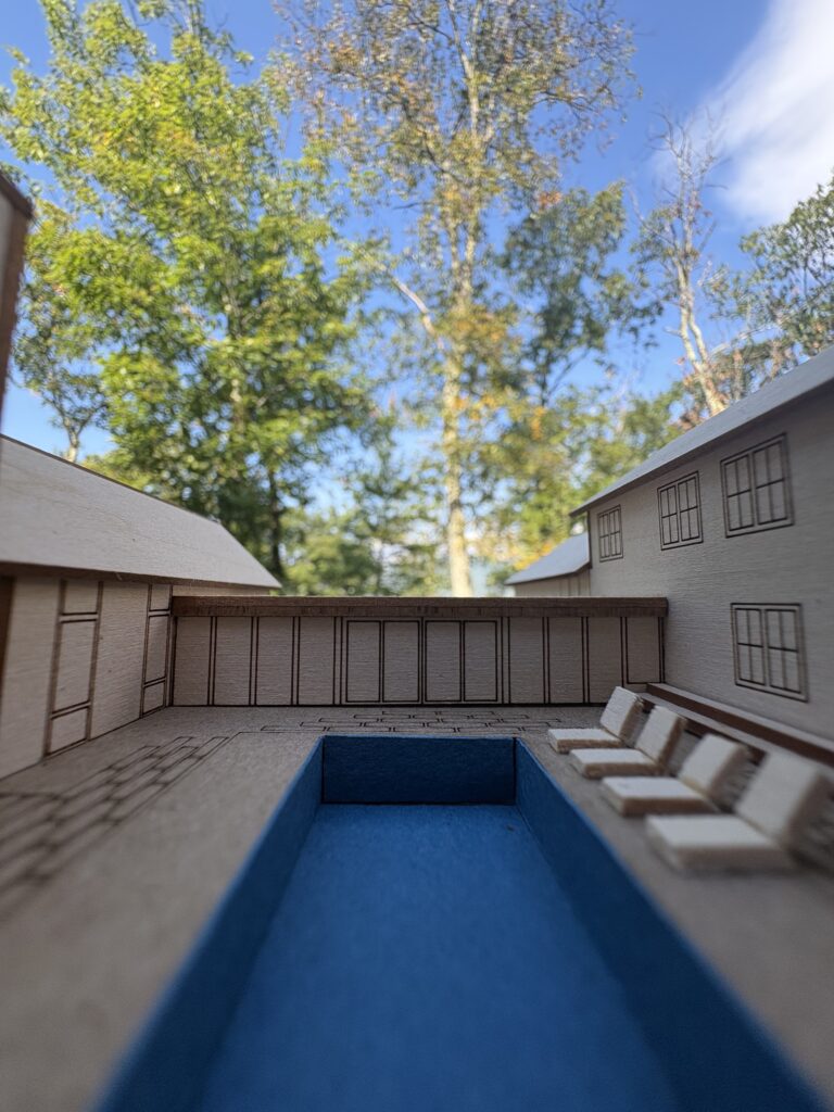 Close-up inside a wood architectural model looking down a blue lap pool lined with tiny lounge chairs, with real trees and sky behind.