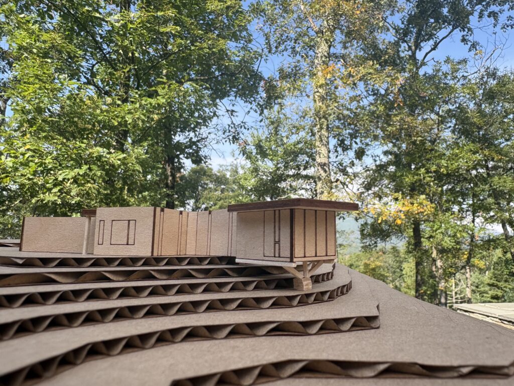 Wood model of a low modern home cantilevered over stacked cardboard contour layers, photographed outdoors against tall trees and distant hills.