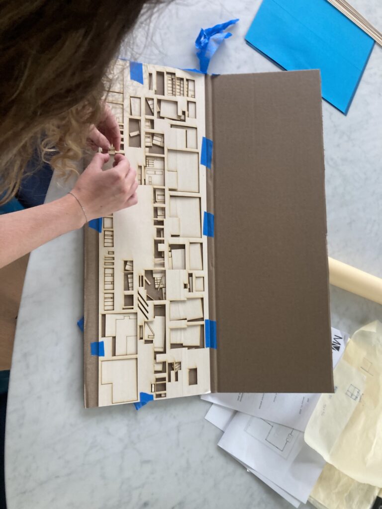 Overhead view of a person lifting small pieces from a laser-cut wood sheet taped to cardboard, with floor plan sketches on the table.