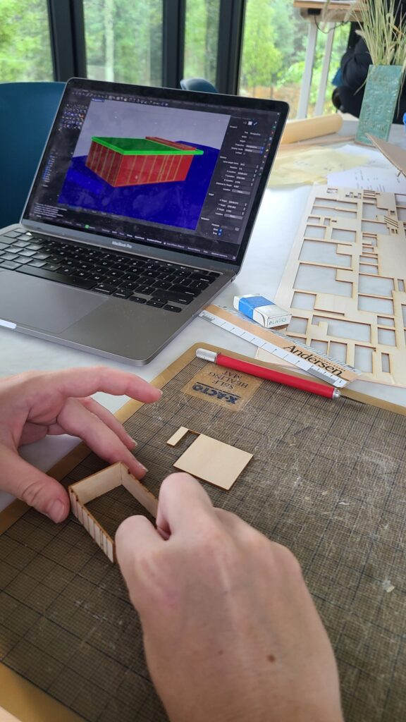 Hands assembling a small laser-cut wood model wall on a cutting mat, with a 3D digital model of the building open on a laptop behind.