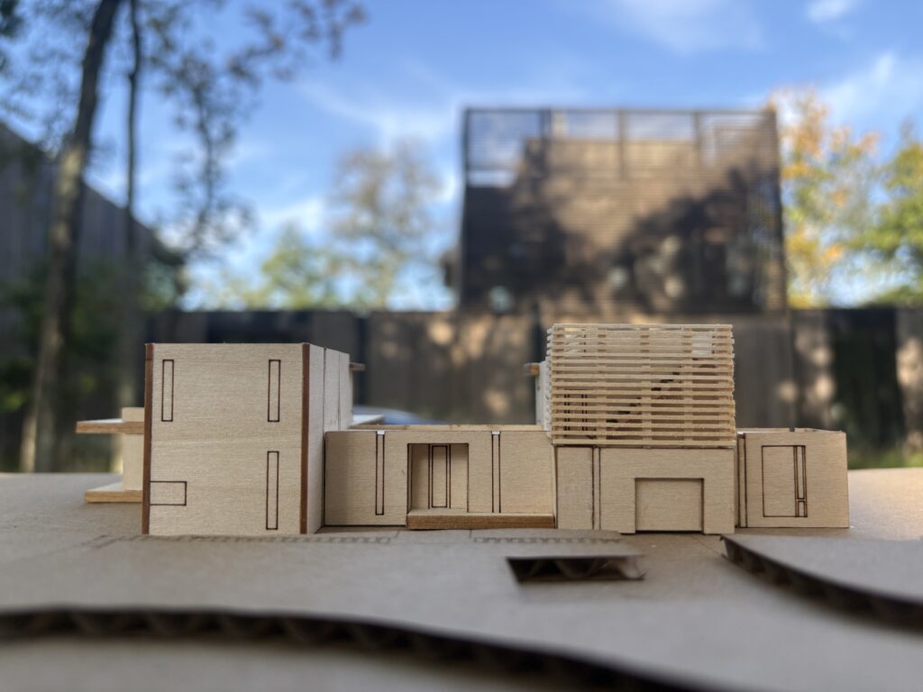 Wood architectural model with a slatted upper-level screen, set in front of the finished dark wood home with the same screened volume.